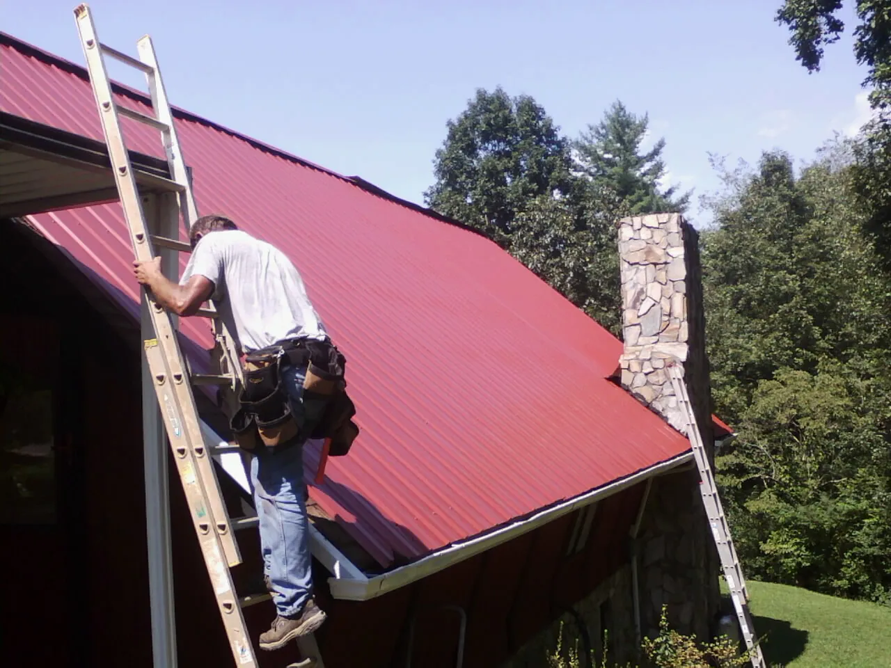 Roofer on a ladder beside a red metal roof and stone chimney — Soffit & Fascia Repair by Roofing Brothers in Slate Run